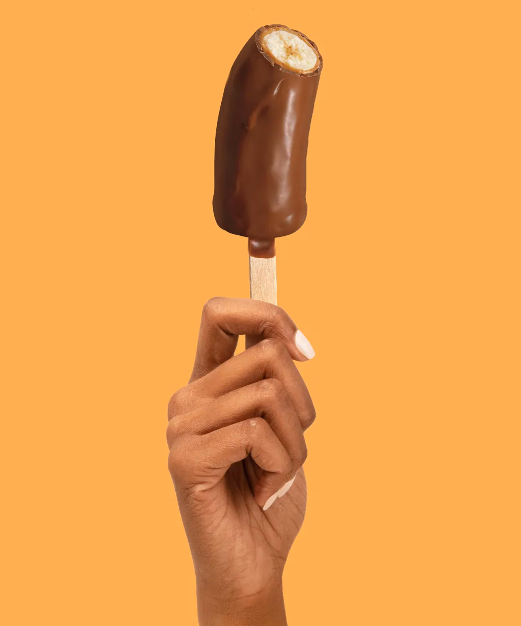 Hand with white nails holding an unwrapped chocolate-dipped frozen banana on a wooden stick on orange.