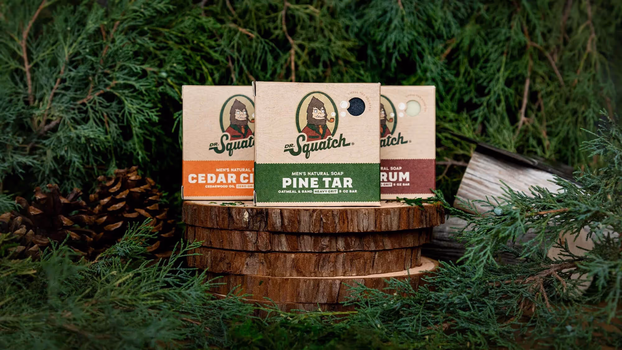Three Dr. Squatch soap boxes including Cedar Citrus and Pine Tar on stacked wood slices with pine cones.