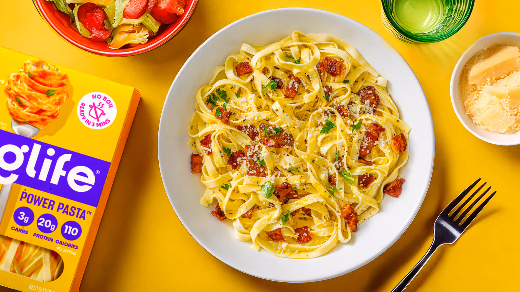 Overhead bowl of fettuccine carbonara on yellow with an egglife Power Pasta box, salad, and grated cheese.
