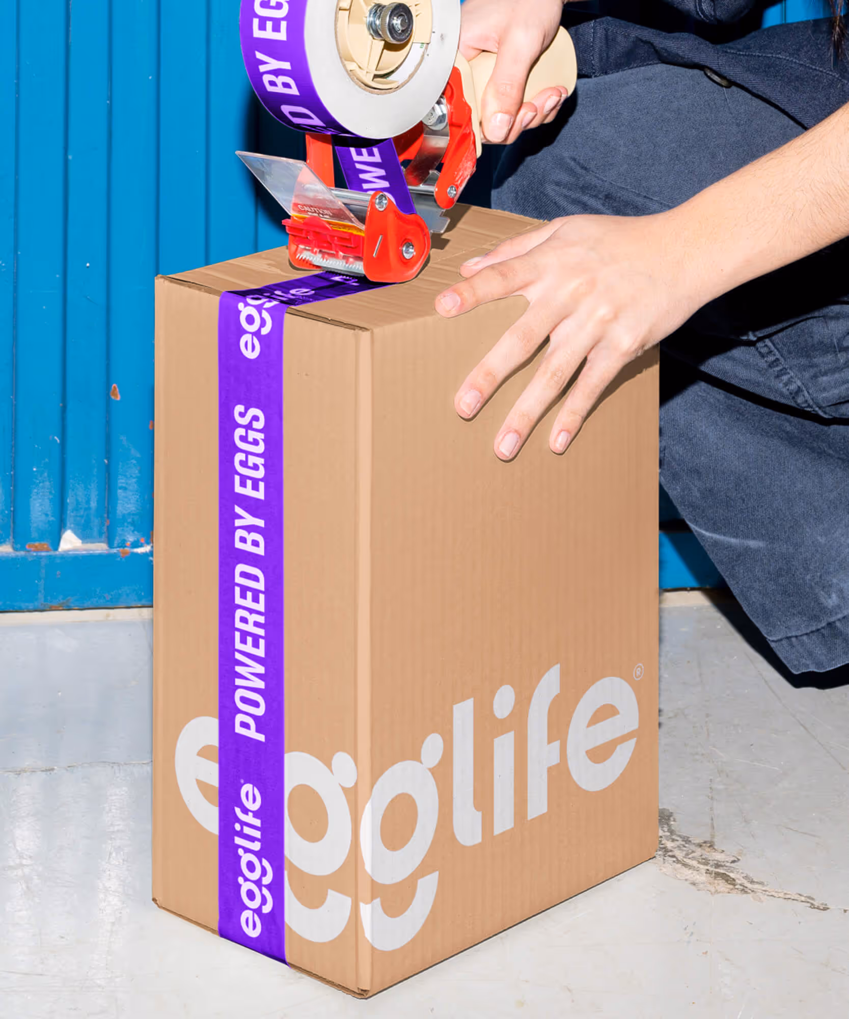 Person sealing a cardboard shipping box with purple 'Powered by eggs' branded tape against a blue garage door.
