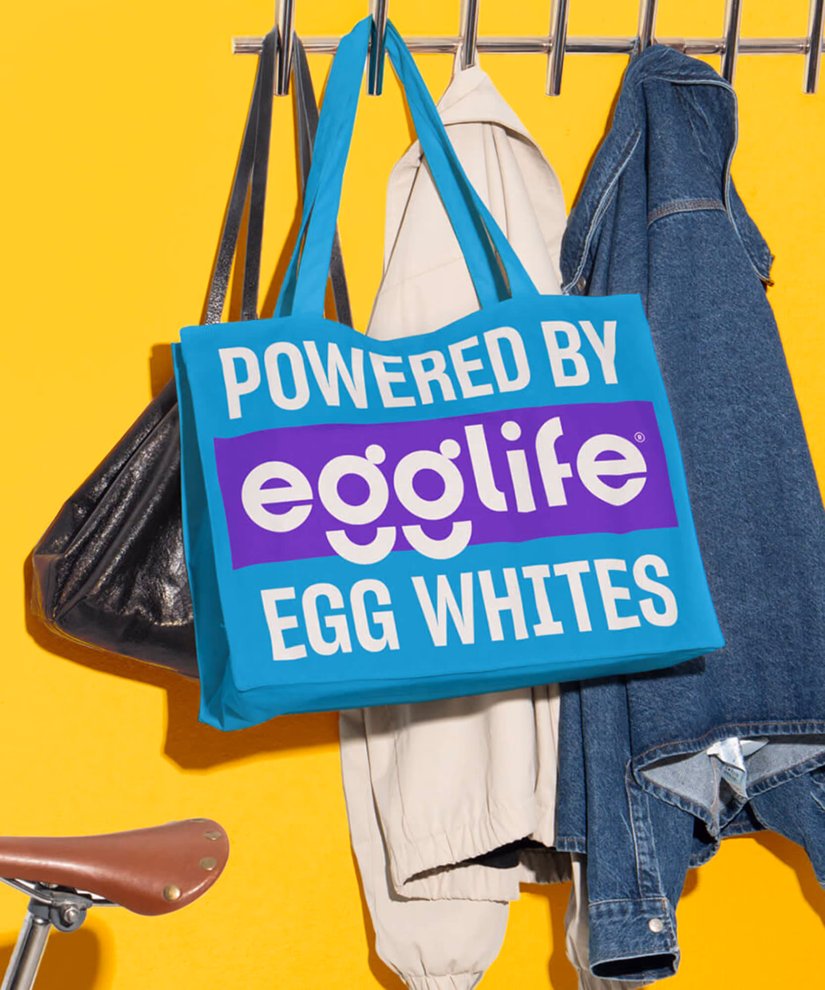 Person in a denim jacket with an orange egglife patch cap; blue 'Powered by egglife egg whites' tote on yellow.