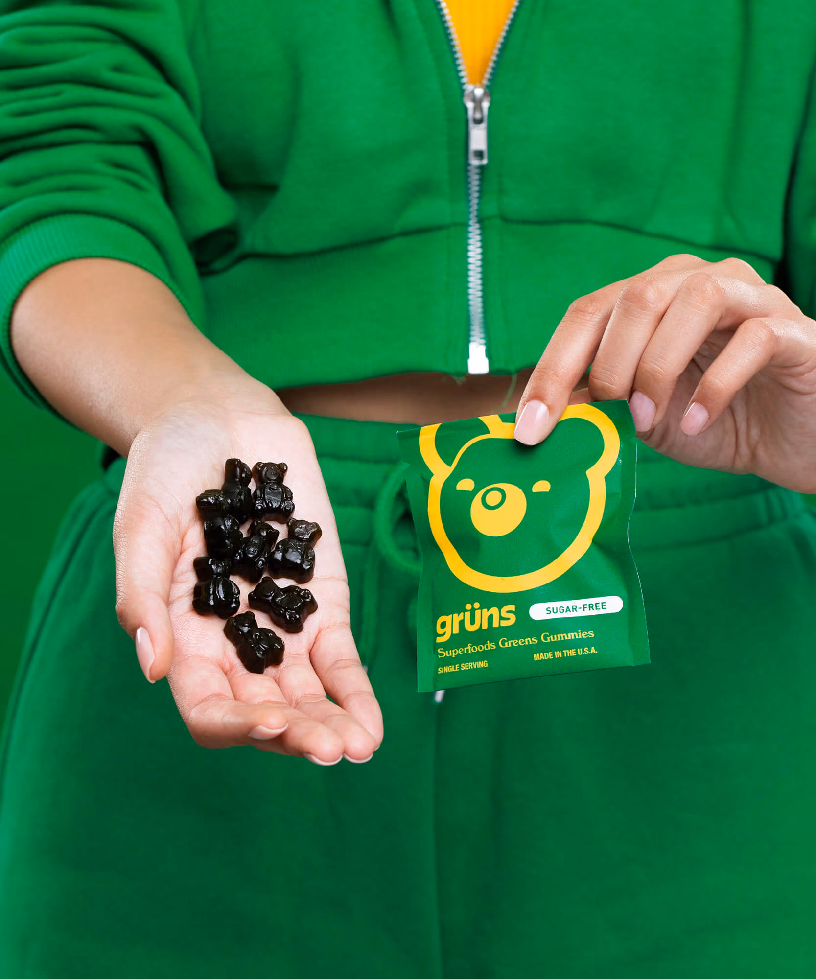 Person in a green hoodie holding dark Grüns gummies and a green Superfoods Greens Gummies sachet with a yellow bear logo.