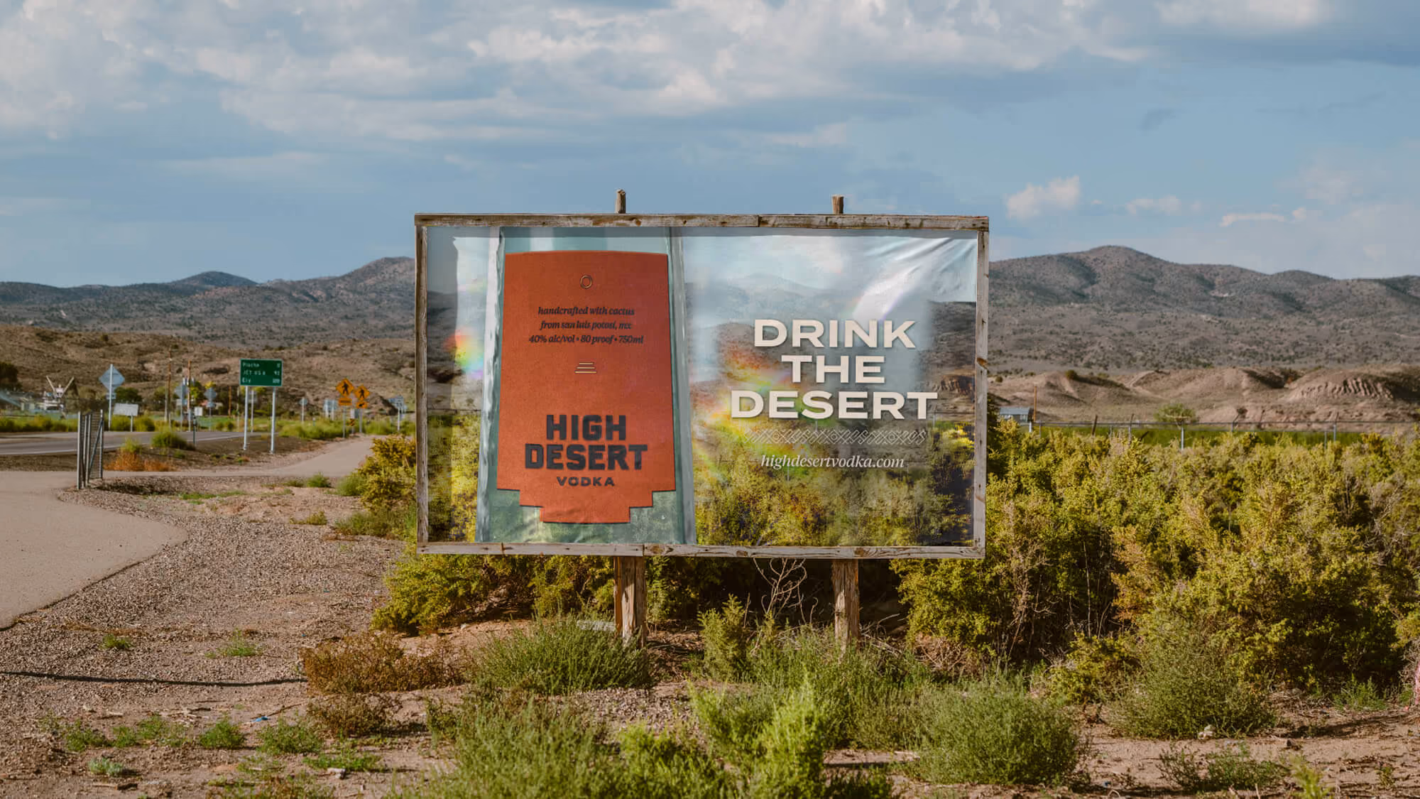 Roadside billboard in arid mountains reading 'Drink the Desert — highdesertvodka.com' beside an orange brand panel.