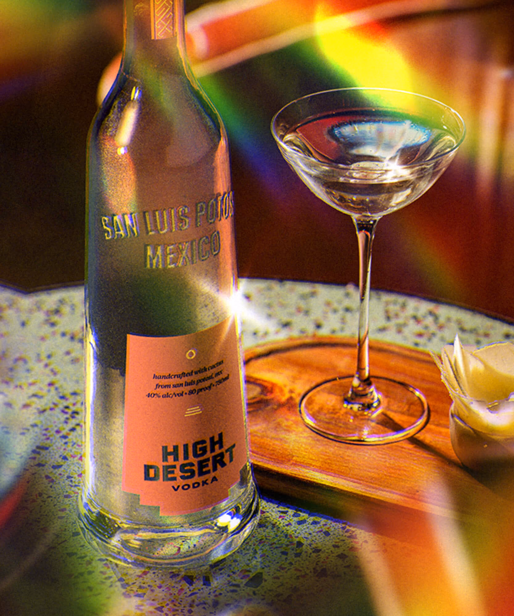High Desert vodka bottle and an empty coupe on a speckled stone tray with prismatic rainbow light streaks.