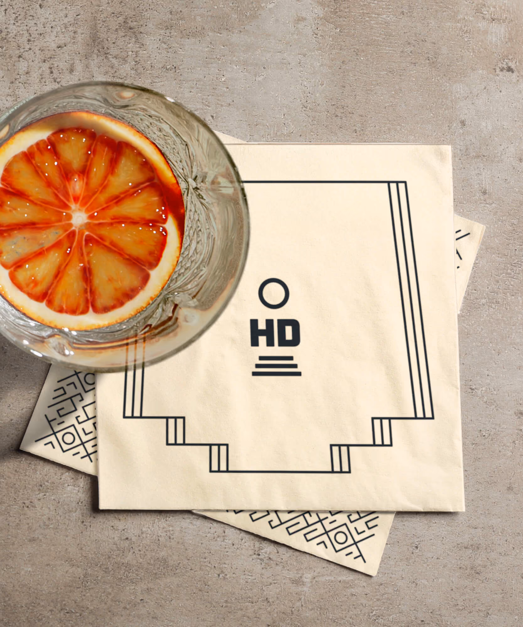 Cream cocktail napkin printed with the stepped 'HD' mark beside a glass with a blood-orange slice on concrete.