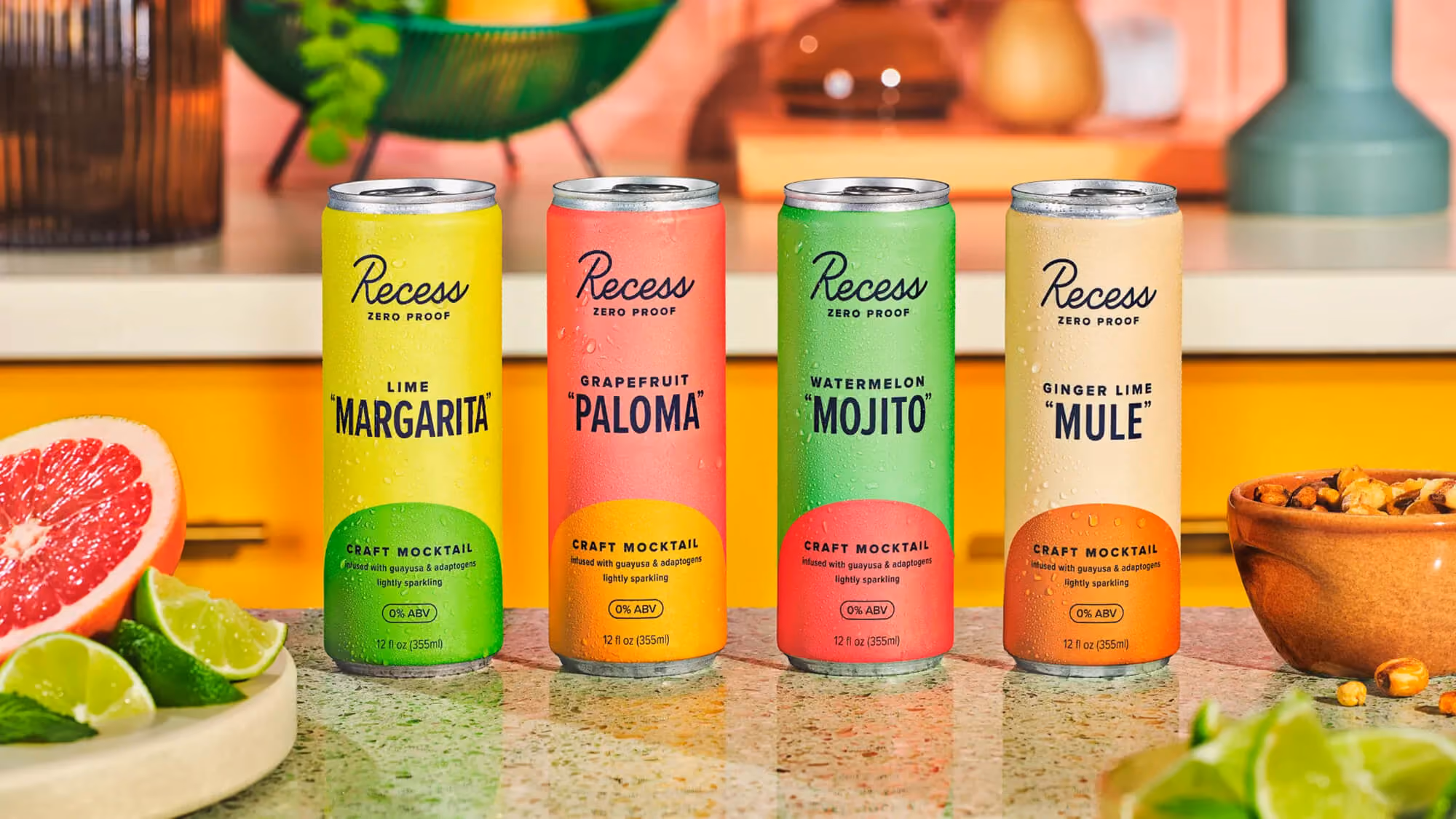 Four Recess Zero Proof cans — Lime Margarita, Grapefruit Paloma, Watermelon Mojito, Ginger Lime Mule — on a counter with citrus fruits and herbs.