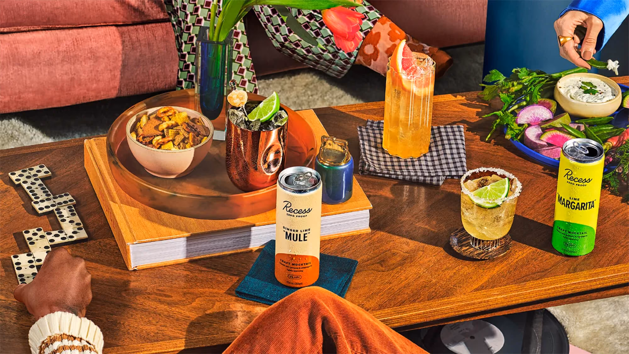 Overhead party table with Recess Zero Proof Ginger Lime Mule and Lime Margarita cans among dominoes, books, snacks, and mixed drinks.