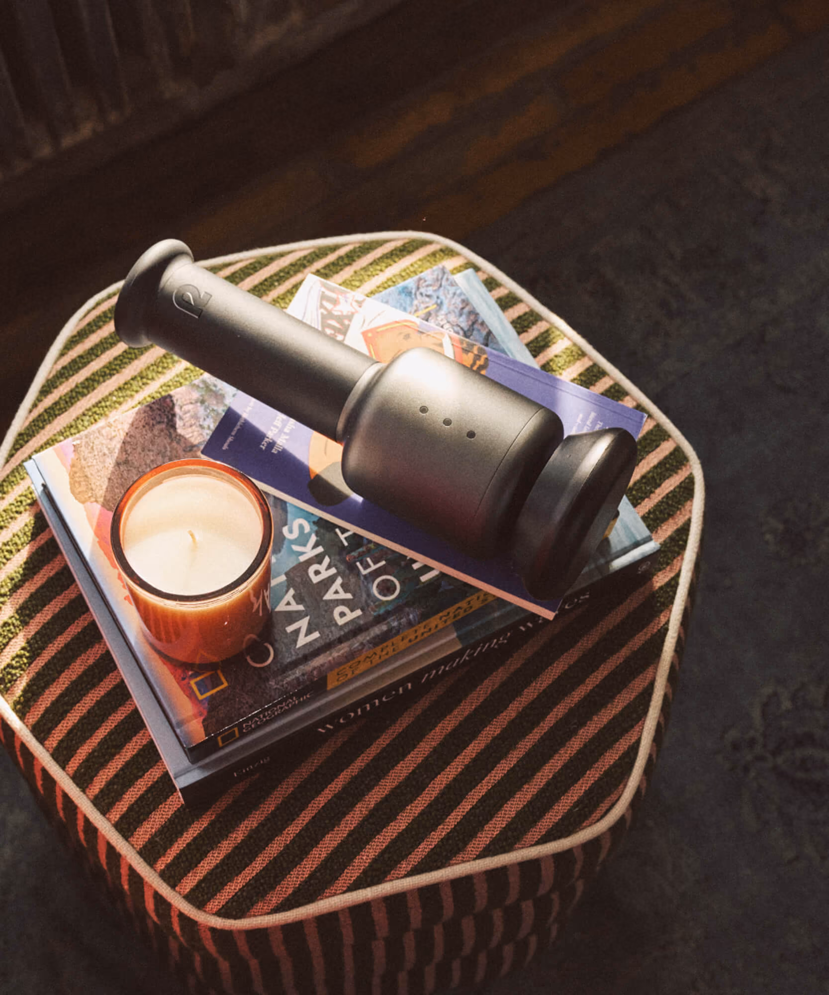 Black Rally massage tool resting on a stack of travel and nature books beside a lit candle on a striped ottoman.