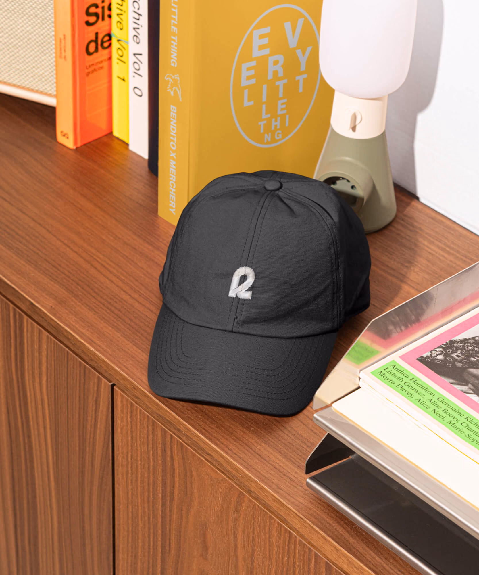 Black Rally baseball cap with embroidered white 'R' on a wooden console with books, lamp, and a yellow poster.