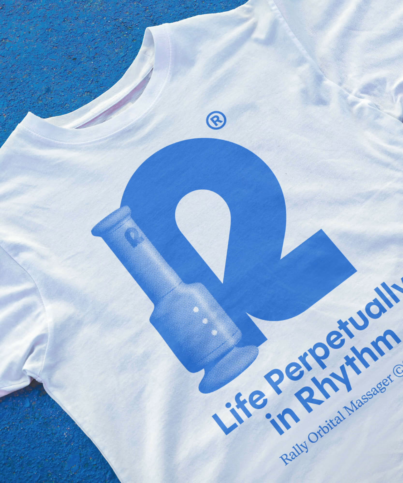 White Rally tee on blue speckled surface printed with a blue 'R' logo and 'Life perpetually in rhythm' tagline.