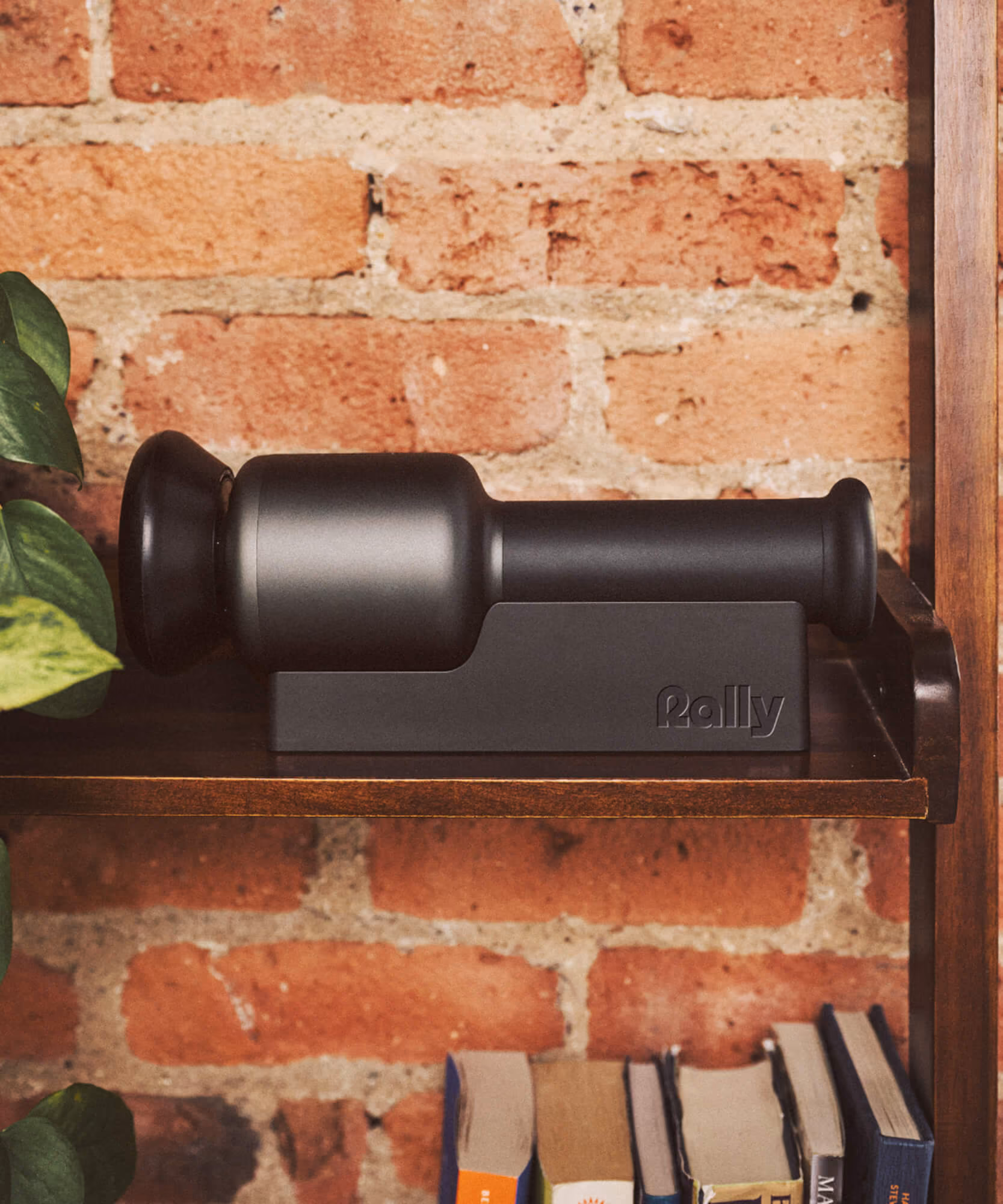 Black Rally massage device docked in its charging base on a wooden shelf against exposed brick with plants and books.