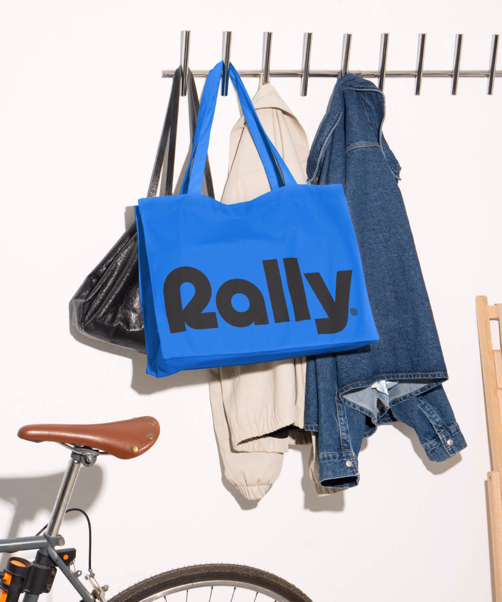 Blue Rally tote bag with the black wordmark hanging on a garment rack with a denim jacket, jeans, and a bicycle below.
