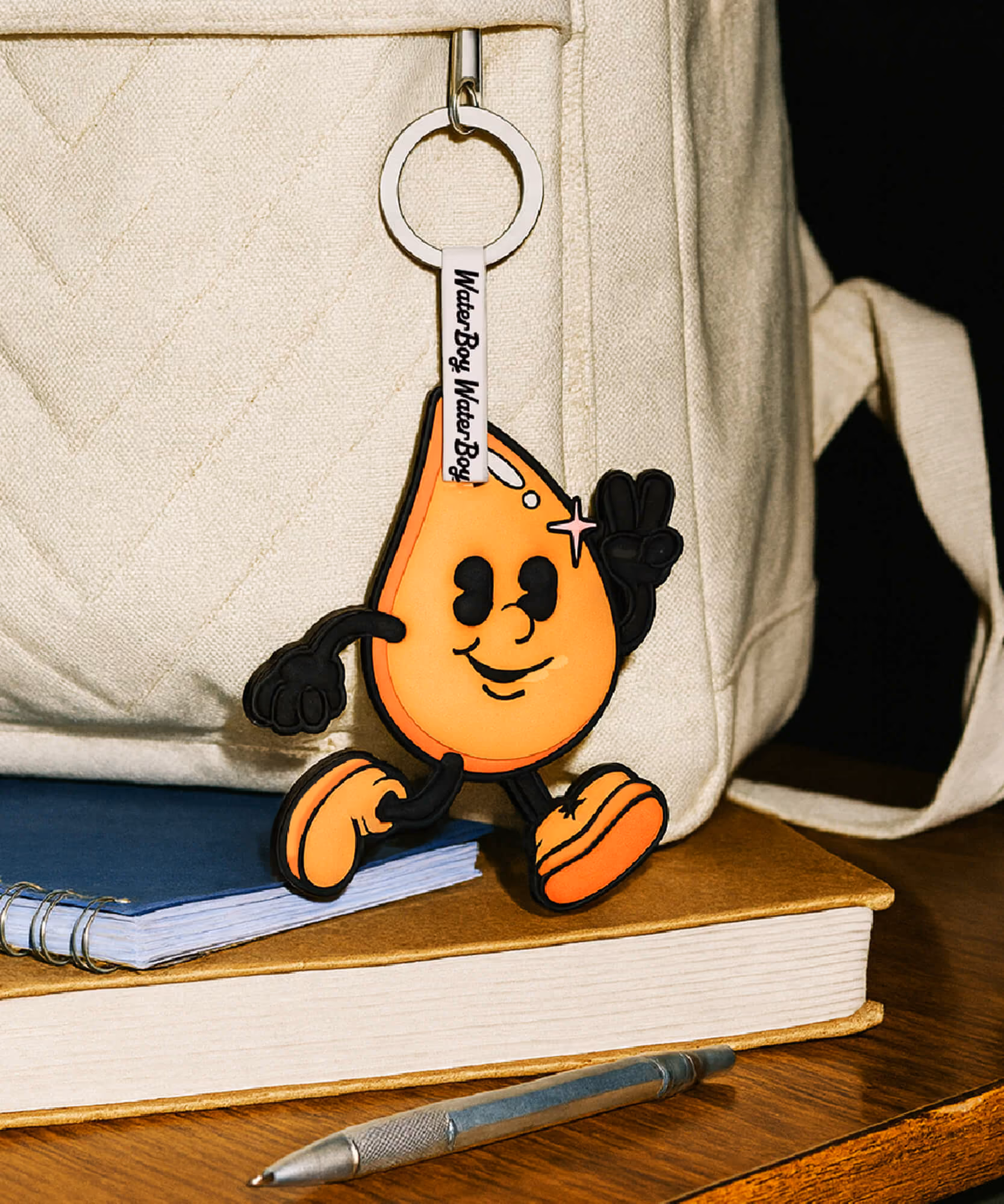A WaterBoy keychain figurine of the orange cartoon drop mascot rests on a quilted bag atop a stack of books.