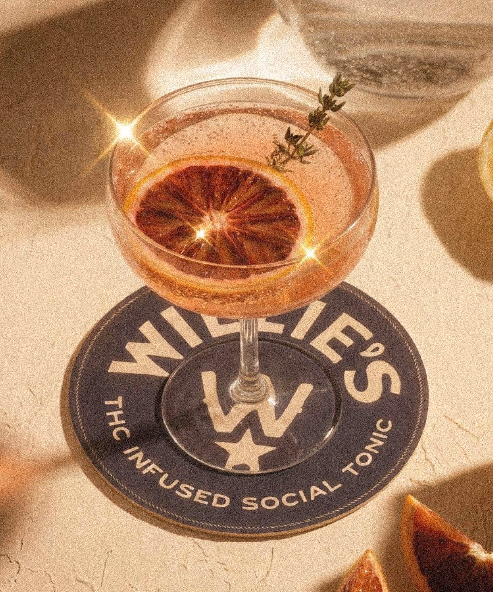 Coupe cocktail garnished with blood orange and thyme on a navy "Willie's THC Infused Social Tonic" coaster