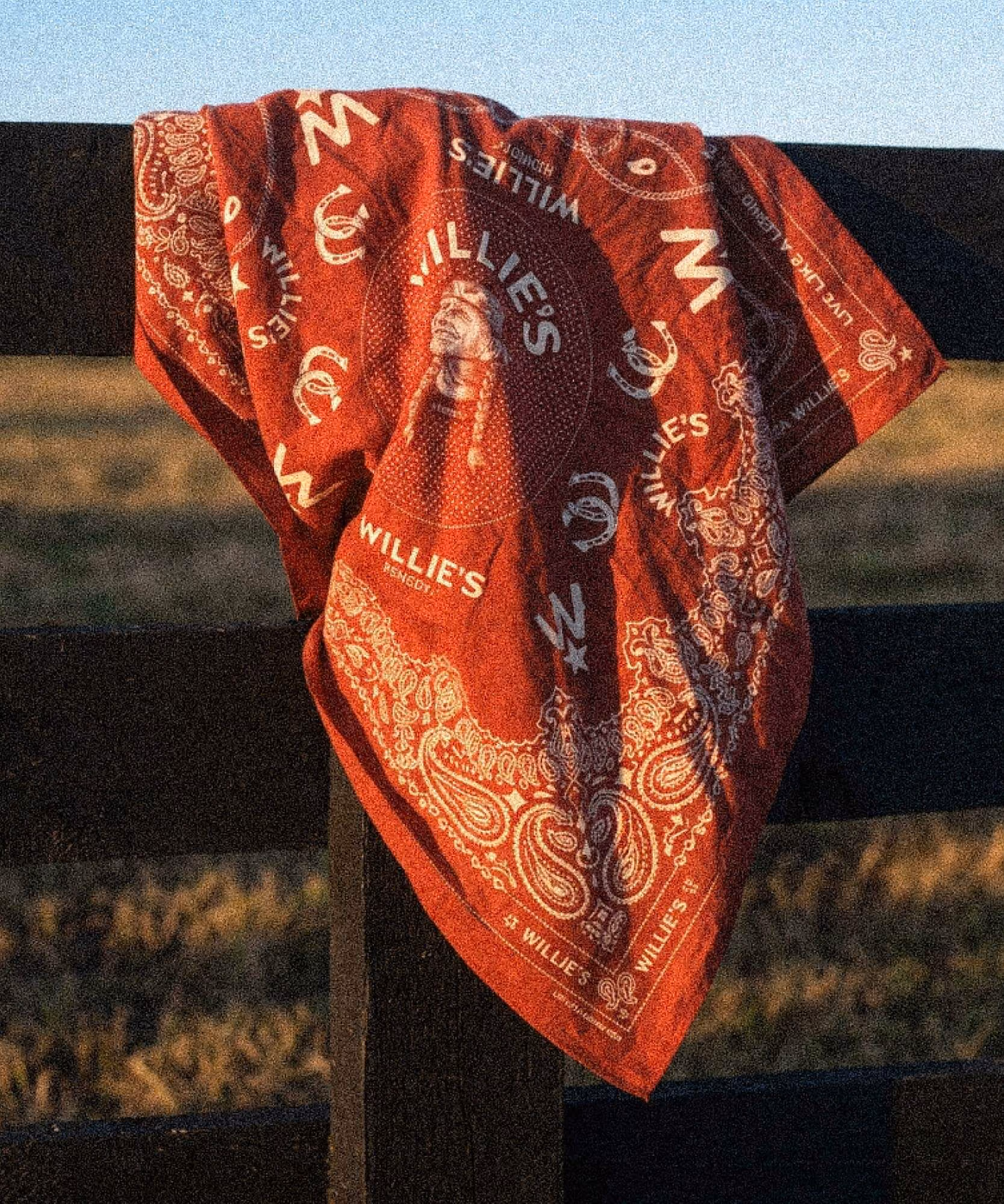 Red paisley bandana printed with Willie's wordmarks, horseshoes, and portrait, draped over a wooden fence