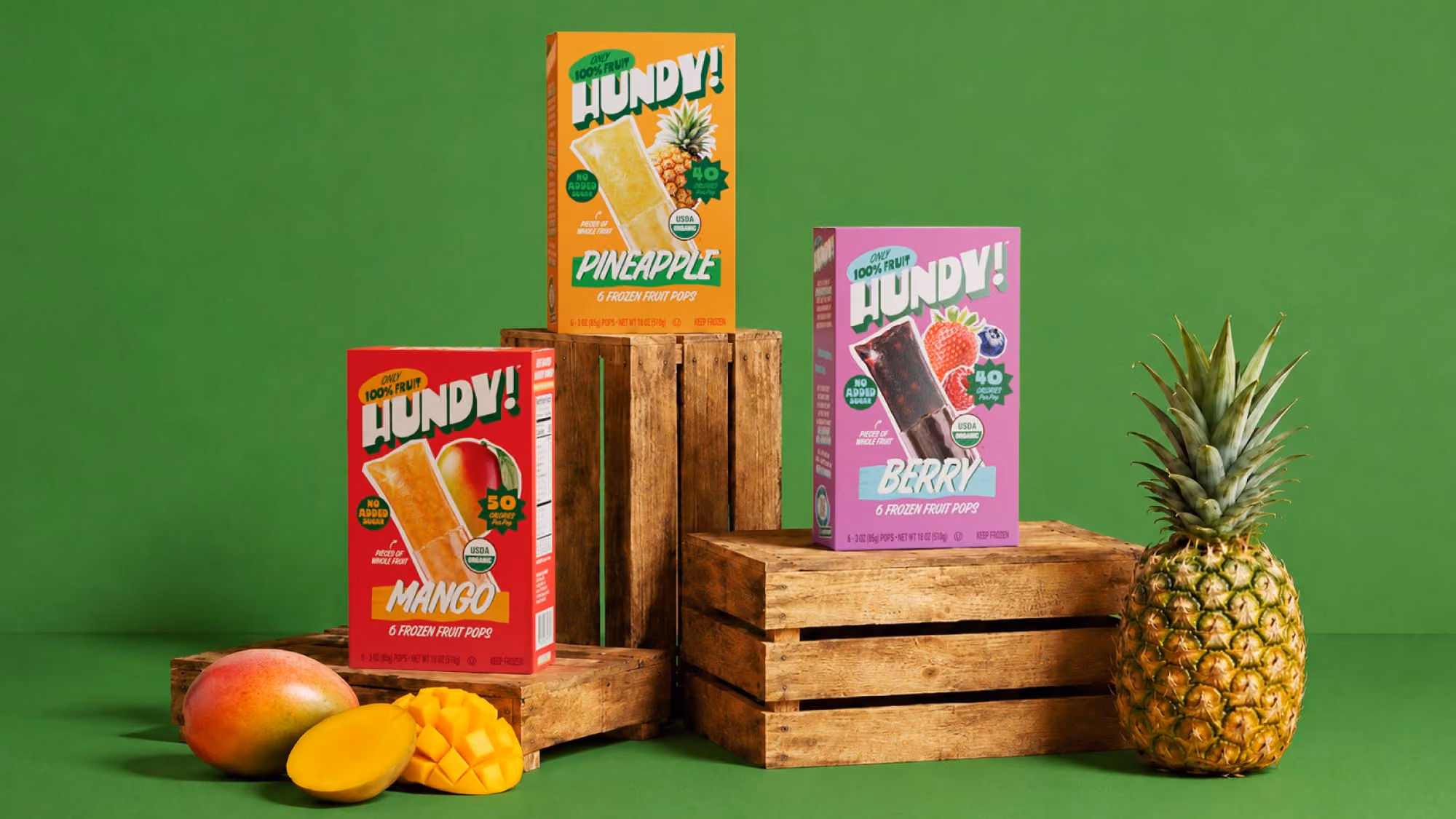 Pineapple, Mango and Berry HUNDY! cartons staged on wooden crates against green with fresh mango and pineapple