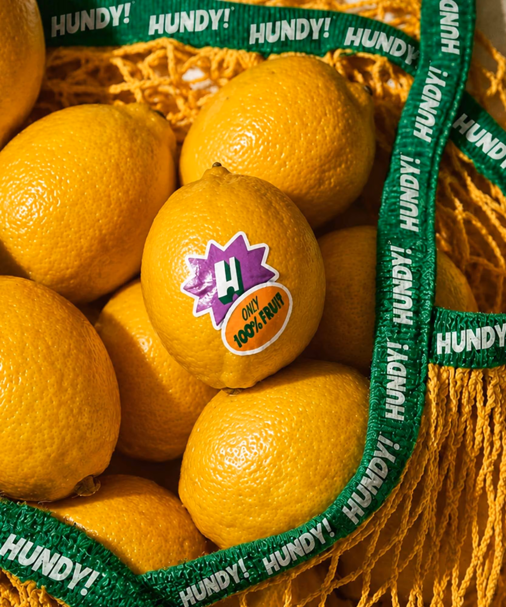 Lemon wearing a purple starburst "H" sticker and orange "ONLY 100% FRUIT" oval in a HUNDY!-branded mesh bag