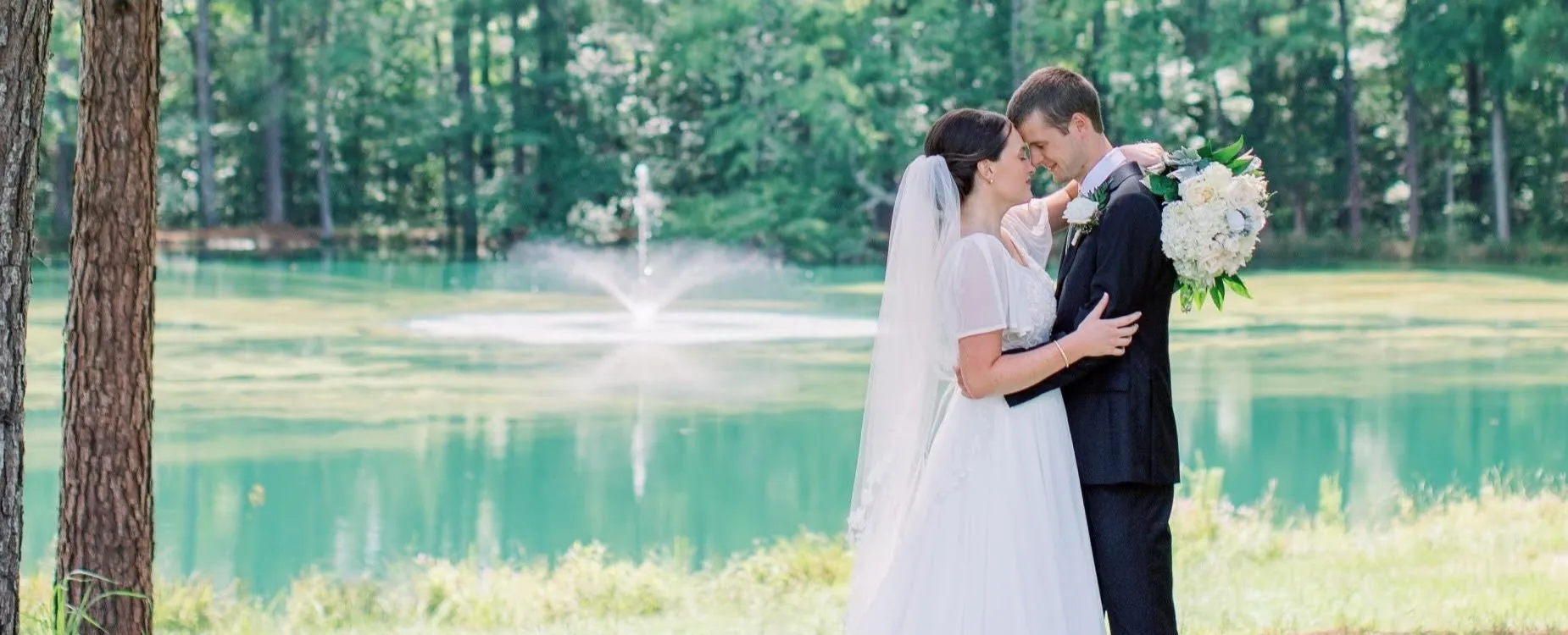 Bride with classic updo and veil embracing groom by a pond with fountain at a Raleigh NC wedding venue, styled by Dayln Faith Hair and Beauty