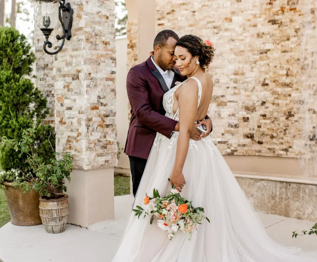 Bride with natural curly updo and coral floral hair accent embracing groom at a stone venue, styled by Dayln Faith Hair and Beauty