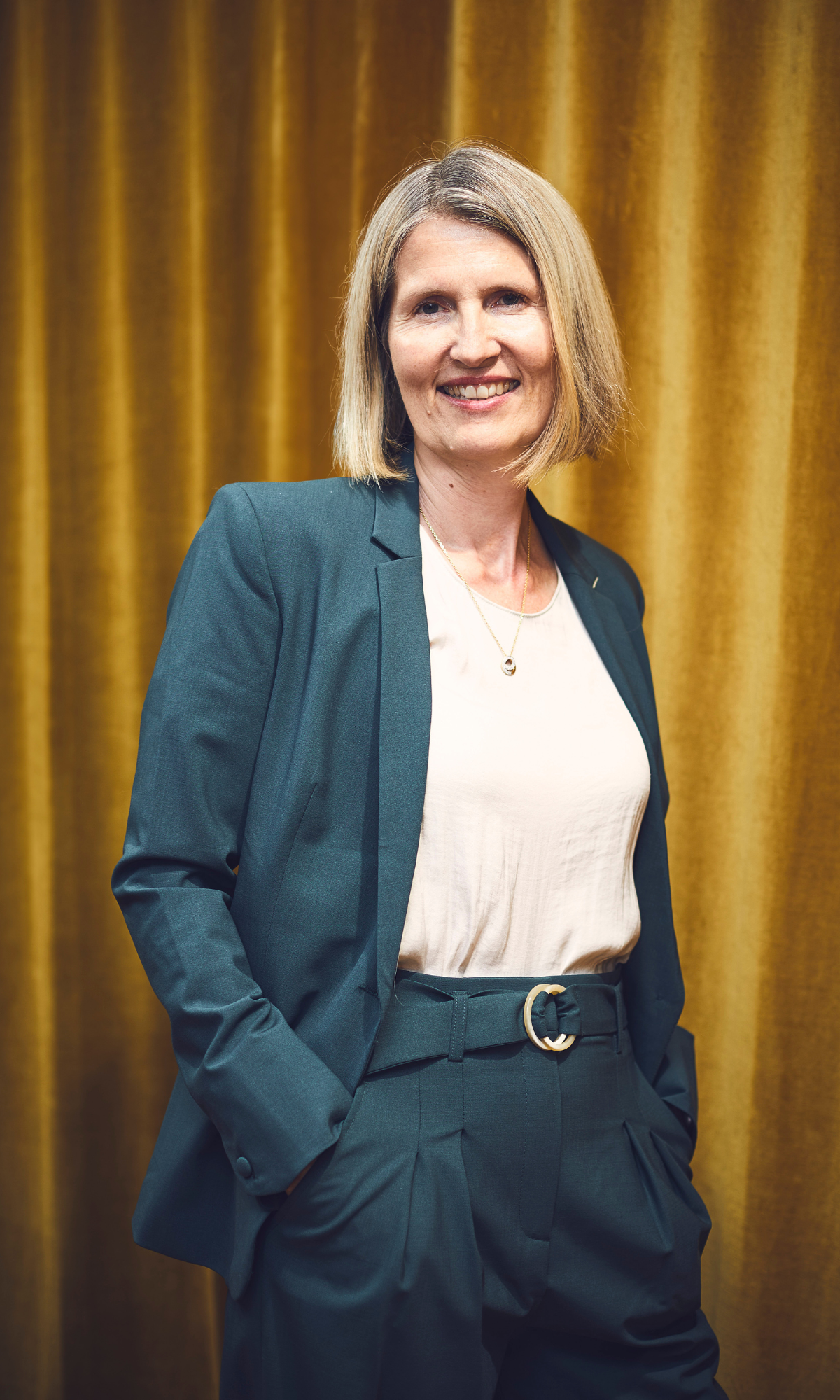 Aurélie, event founder, posing confidently in a teal blazer in front of a gold curtain.
