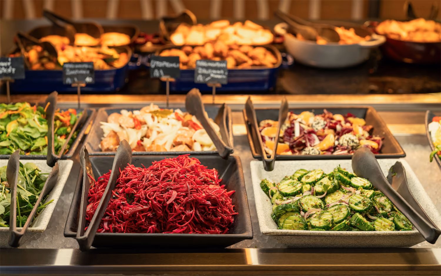Buffet salad bar with dishes of red shredded beets, sliced cucumbers, greens, and mixed salads with tongs.