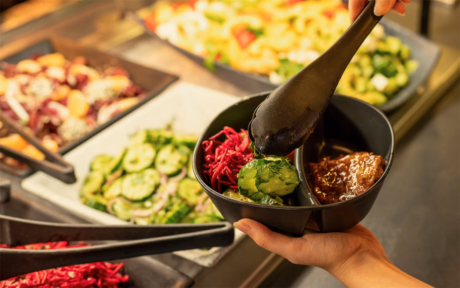Person serving cucumber salad and other dishes into a black divided bowl with tongs.
