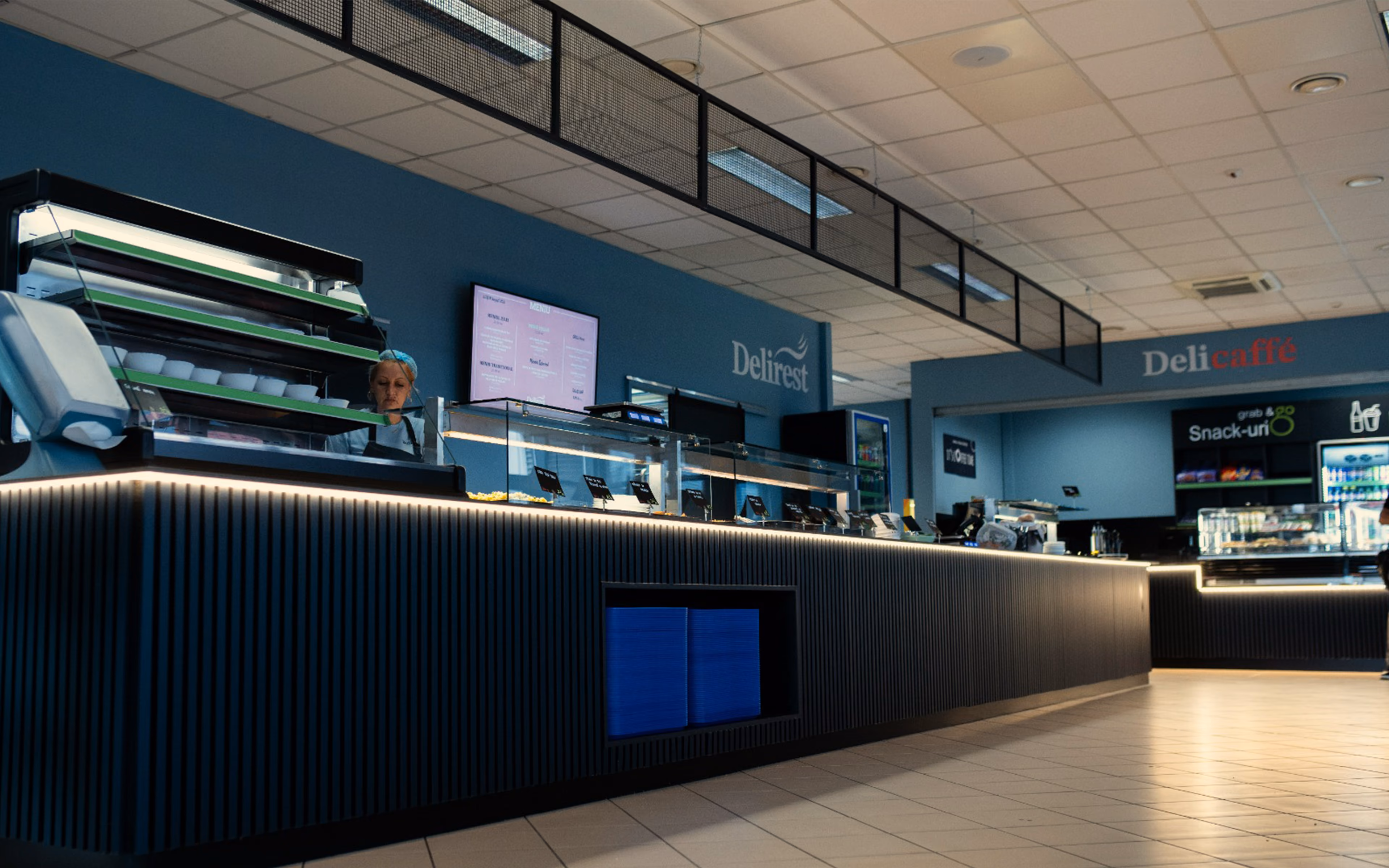 Modern delicatessen counter with a staff member, glass food displays, and menu screens on blue walls.