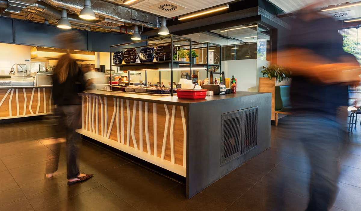 Modern cafeteria counter with food trays and people walking by in motion blur.