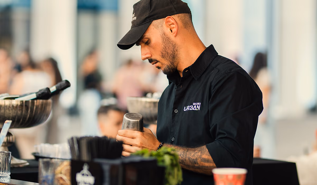 Man in a black uniform and cap preparing a cocktail at a bar with blurred crowd behind him.