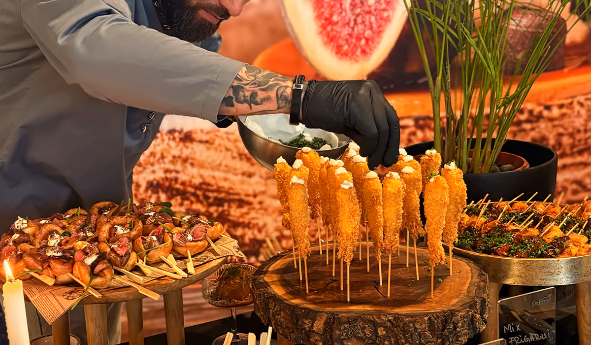 Chef wearing black gloves garnishing breaded appetizers on wooden skewers at a catering station.