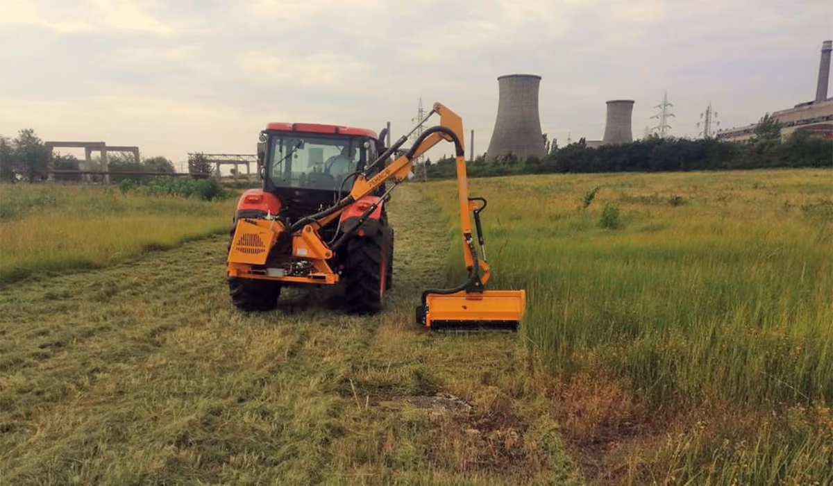 Red tractor with an orange mower cutting grass in a field near an industrial power plant.