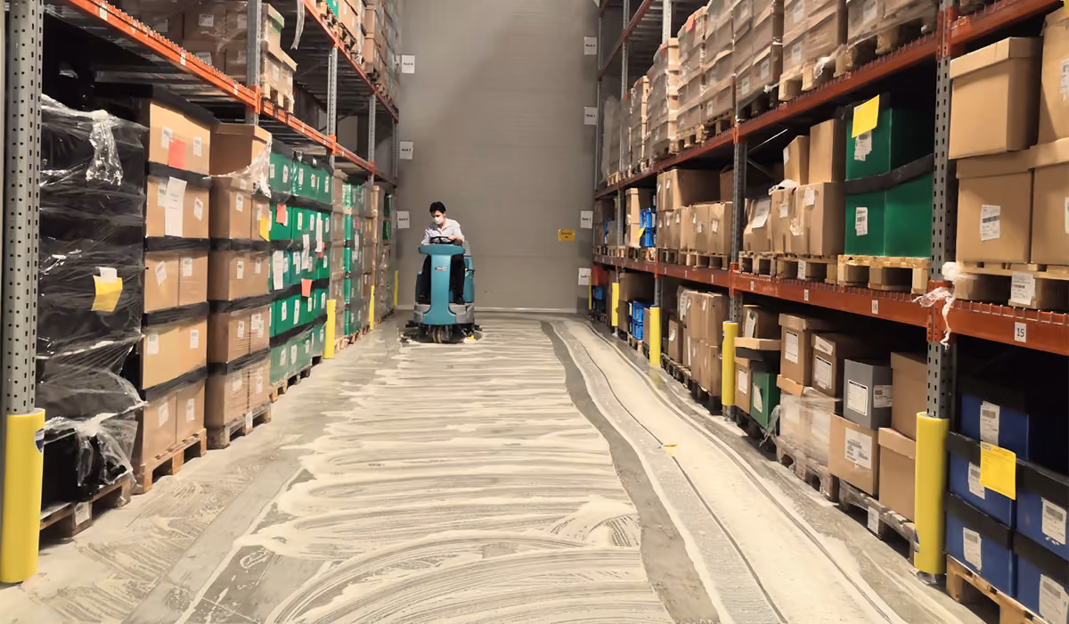 Person on floor cleaning machine cleaning aisle between warehouse shelves with stacked boxes and pallets.