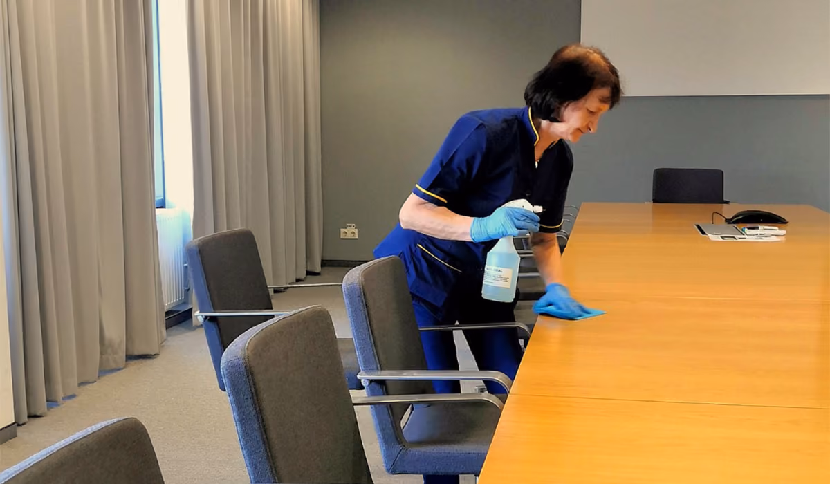 Person wearing gloves cleaning a large conference room table with spray bottle and cloth.