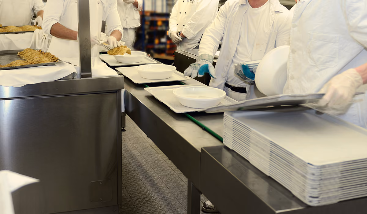 Chefs wearing white uniforms serving food on plates along a stainless steel kitchen counter.