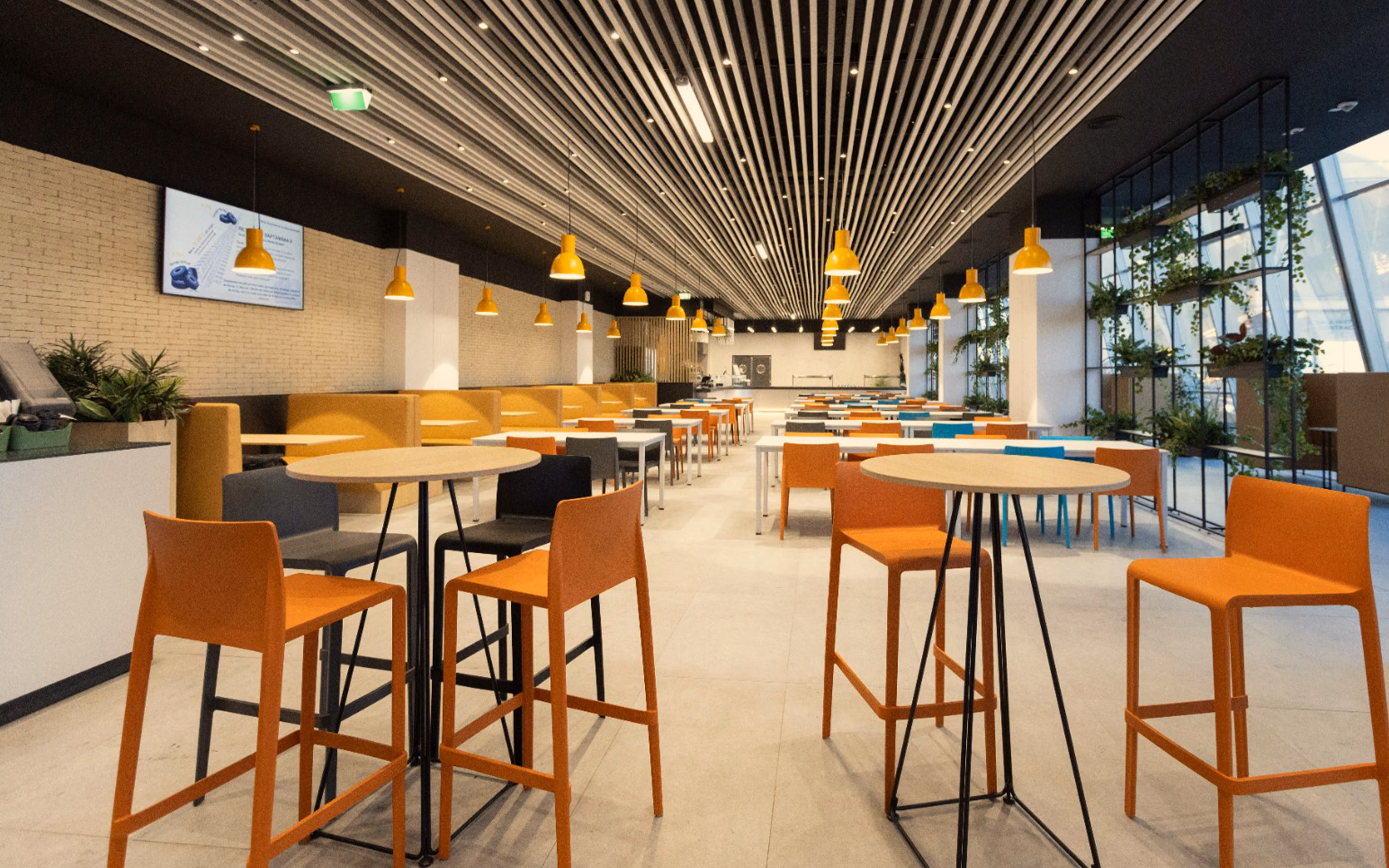Modern cafeteria with orange and gray chairs, round tables, yellow pendant lights, and plants along windows.