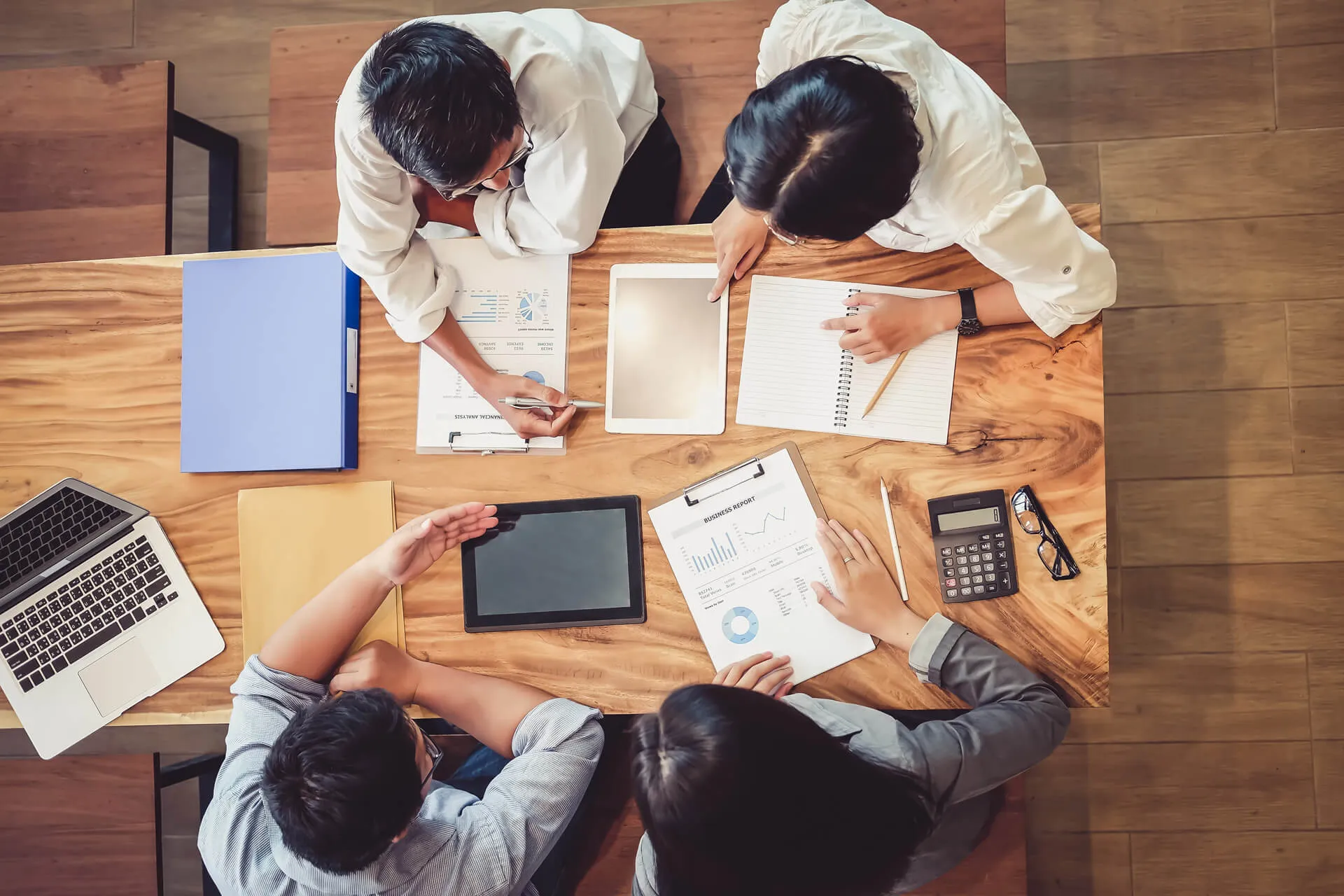 Four people sitting at a wooden table reviewing charts, reports, and digital tablets in a meeting.