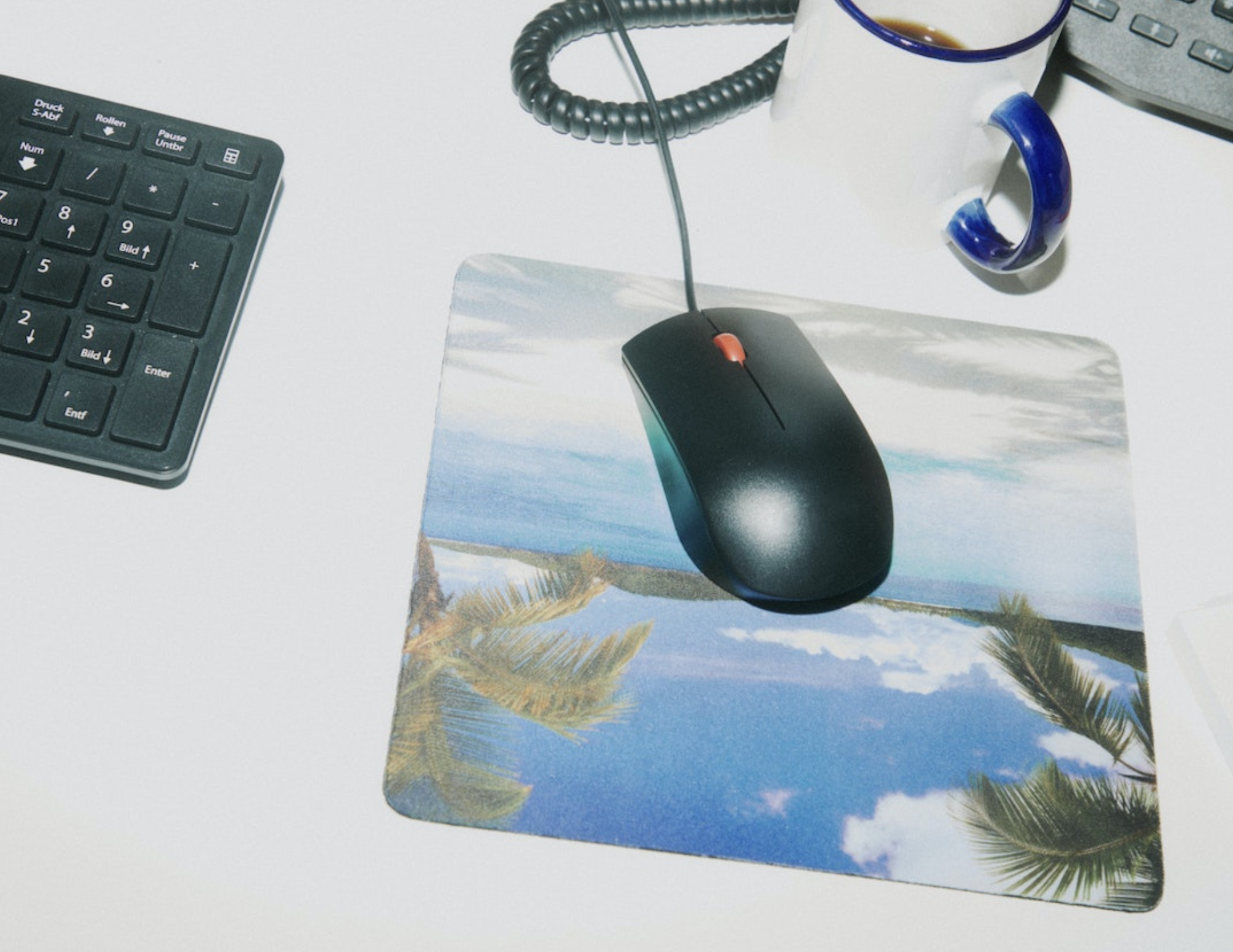 Computer mouse on a tropical beach mouse pad beside a keyboard and coffee mug