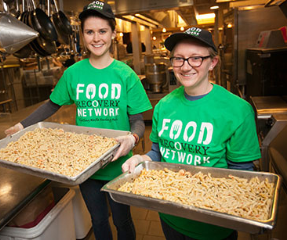 Two Food Recovery Network volunteers holding trays of pasta in a commercial kitchen