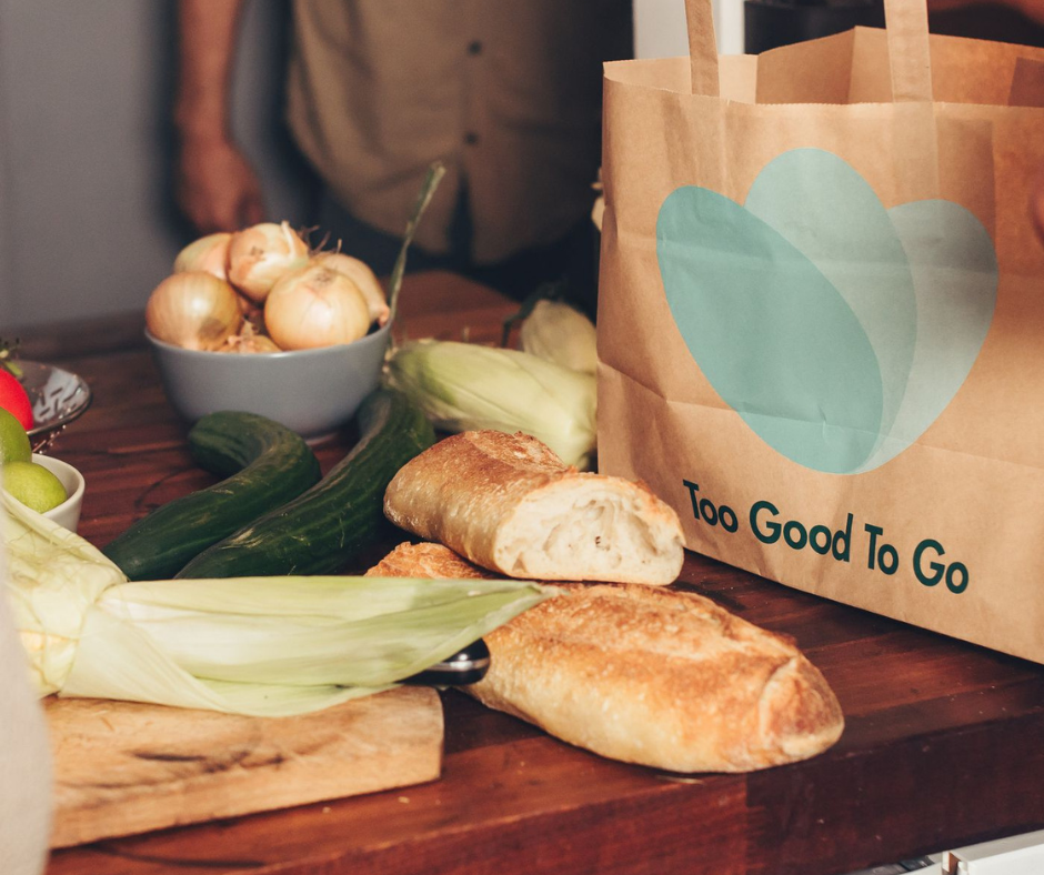 Too Good To Go paper bag beside bread, corn and vegetables on a kitchen counter