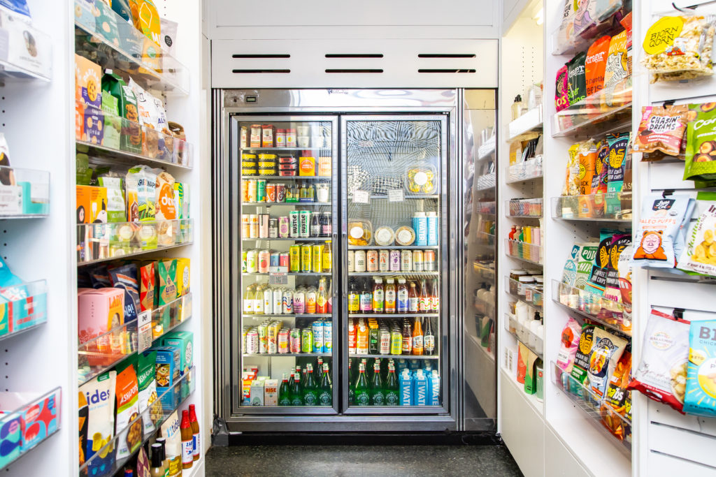 Narrow aisle at The Goods Mart lined with snacks beside a glass-door drinks cooler
