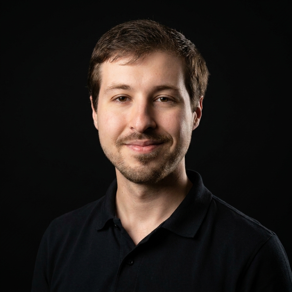 Portrait of a smiling young man with short brown hair and light facial hair wearing a black polo shirt against a black background.