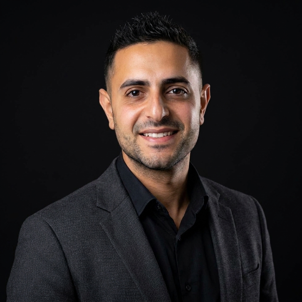 Smiling man with short dark hair wearing a black shirt and dark gray blazer against a black background.