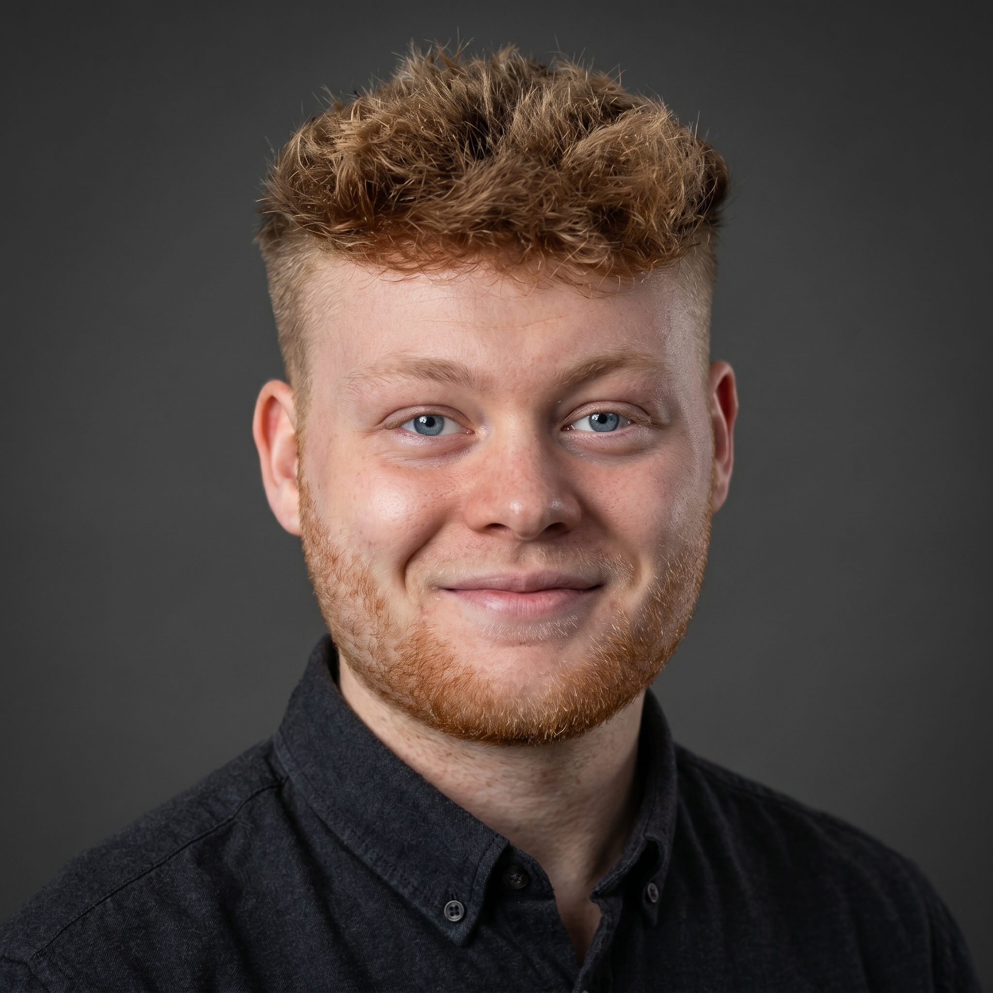 Portrait of a young man with curly red hair and beard, smiling slightly, wearing a gray collared shirt against a black background.