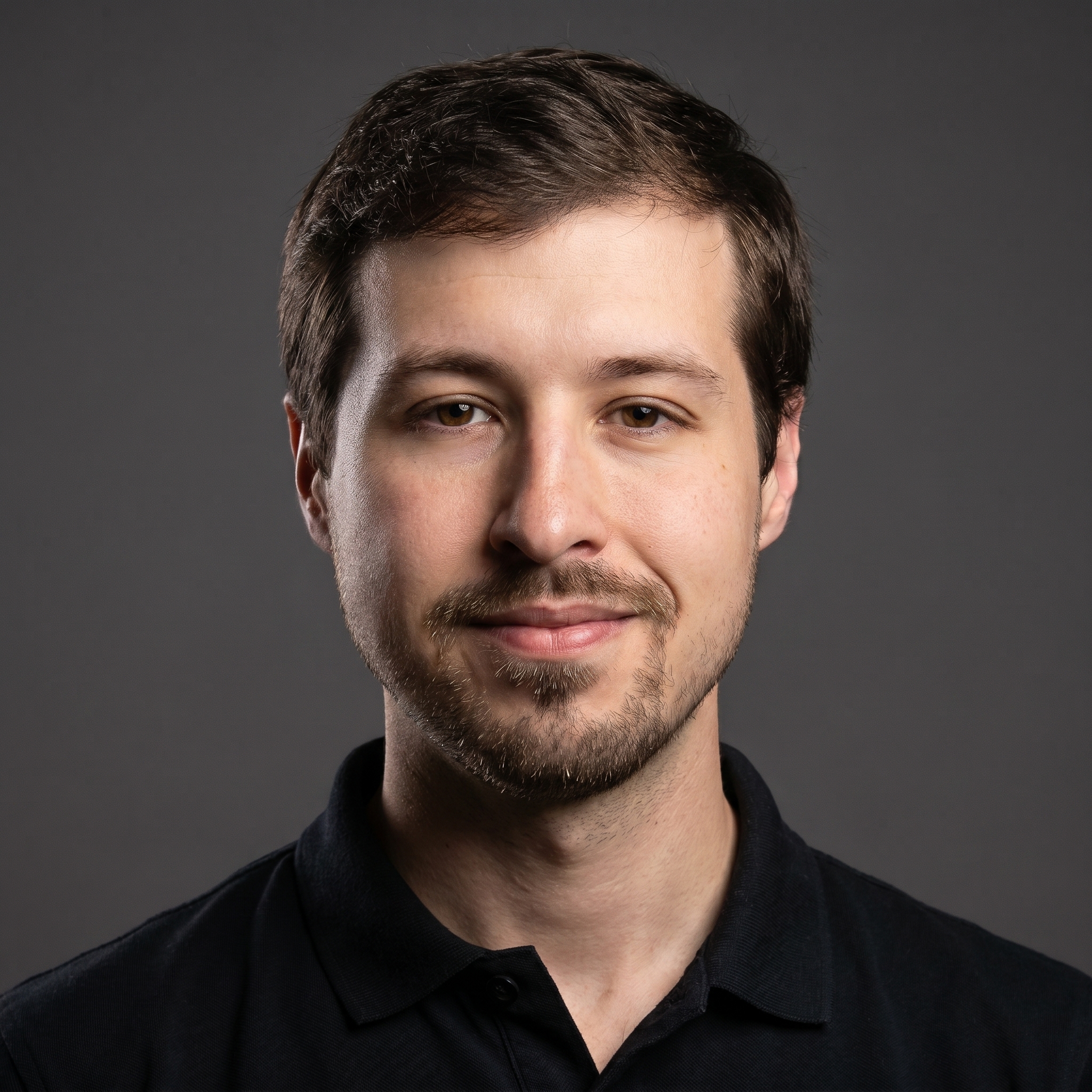 Portrait of a smiling young man with short brown hair and light facial hair wearing a black polo shirt against a black background.