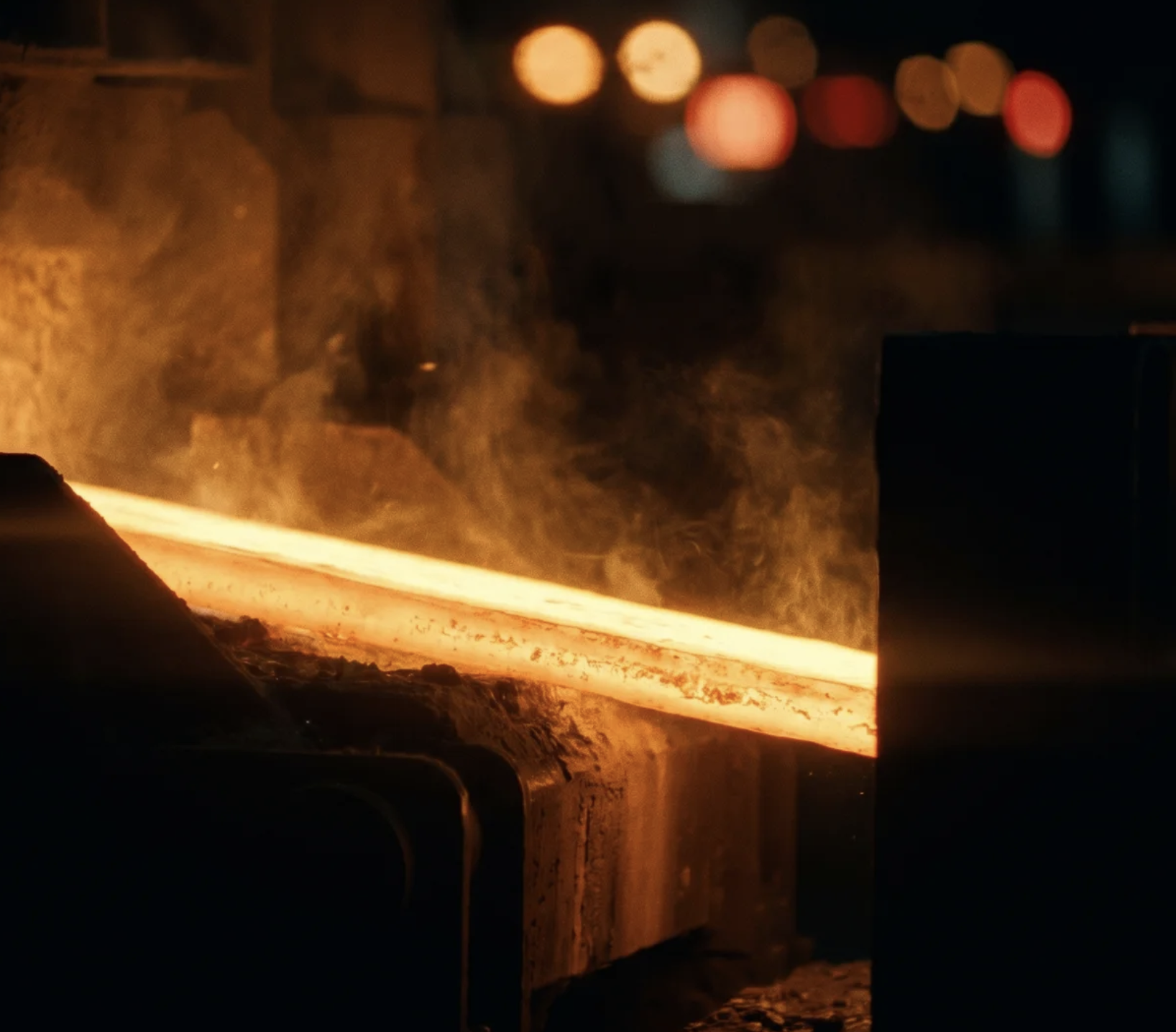 Glowing hot metal bar cooling on industrial equipment with steam rising in a dimly lit factory.