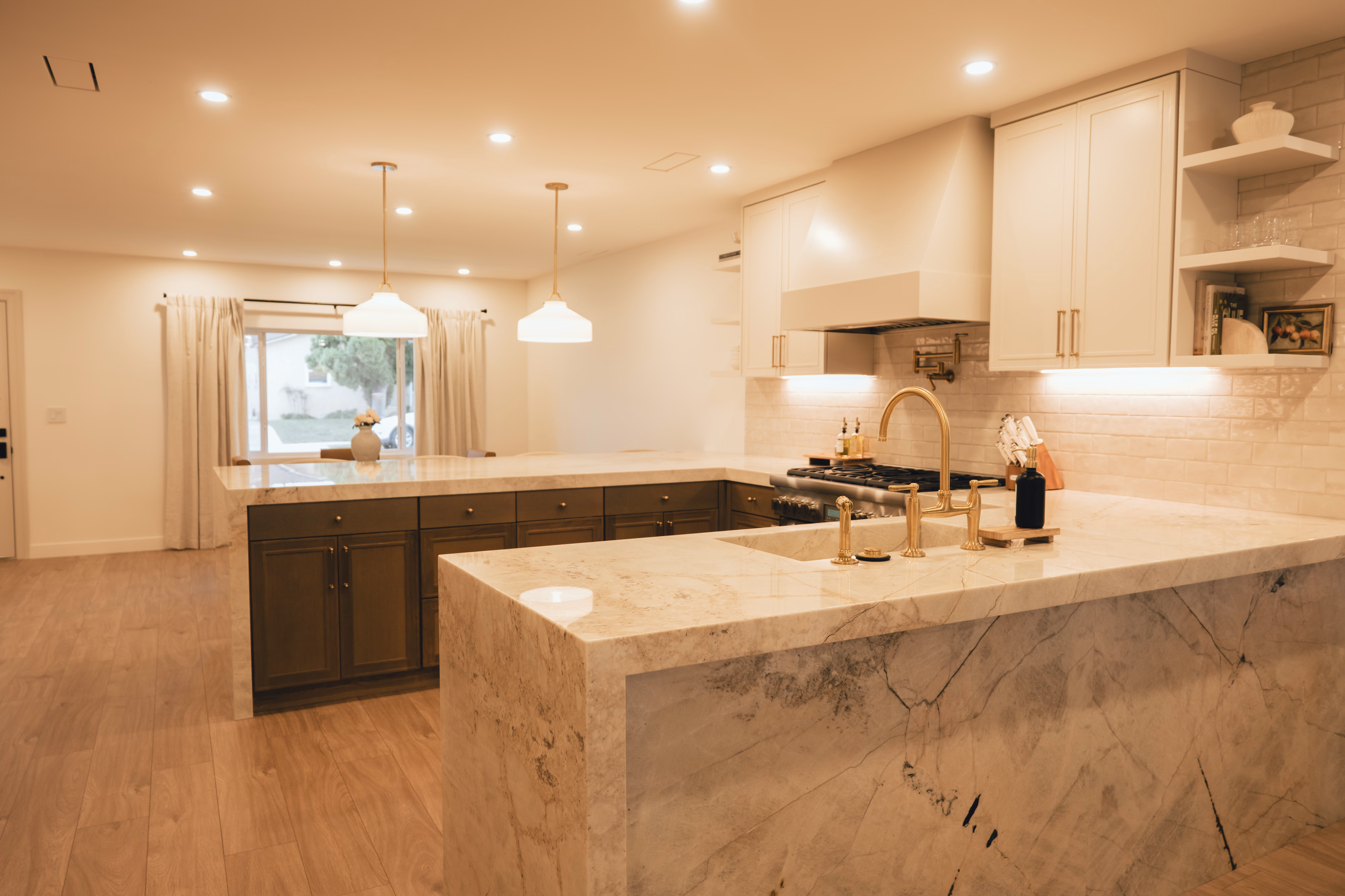 Modern kitchen with marble countertops, brass faucet, dark cabinets, pendant lights, and a large window with curtains.