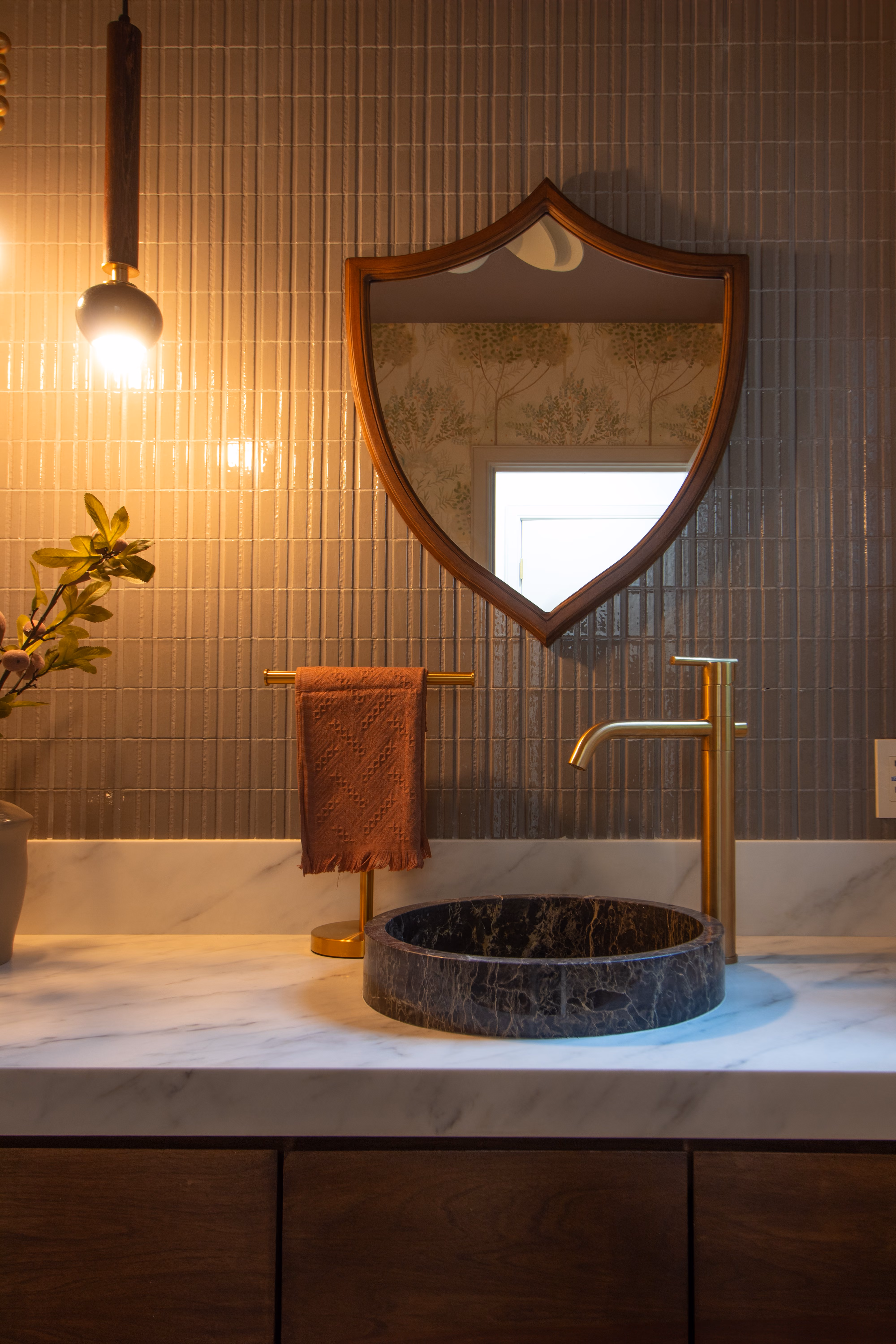 Bathroom vanity with round dark stone sink, gold faucet, towel holder, wooden mirror, and hanging light.