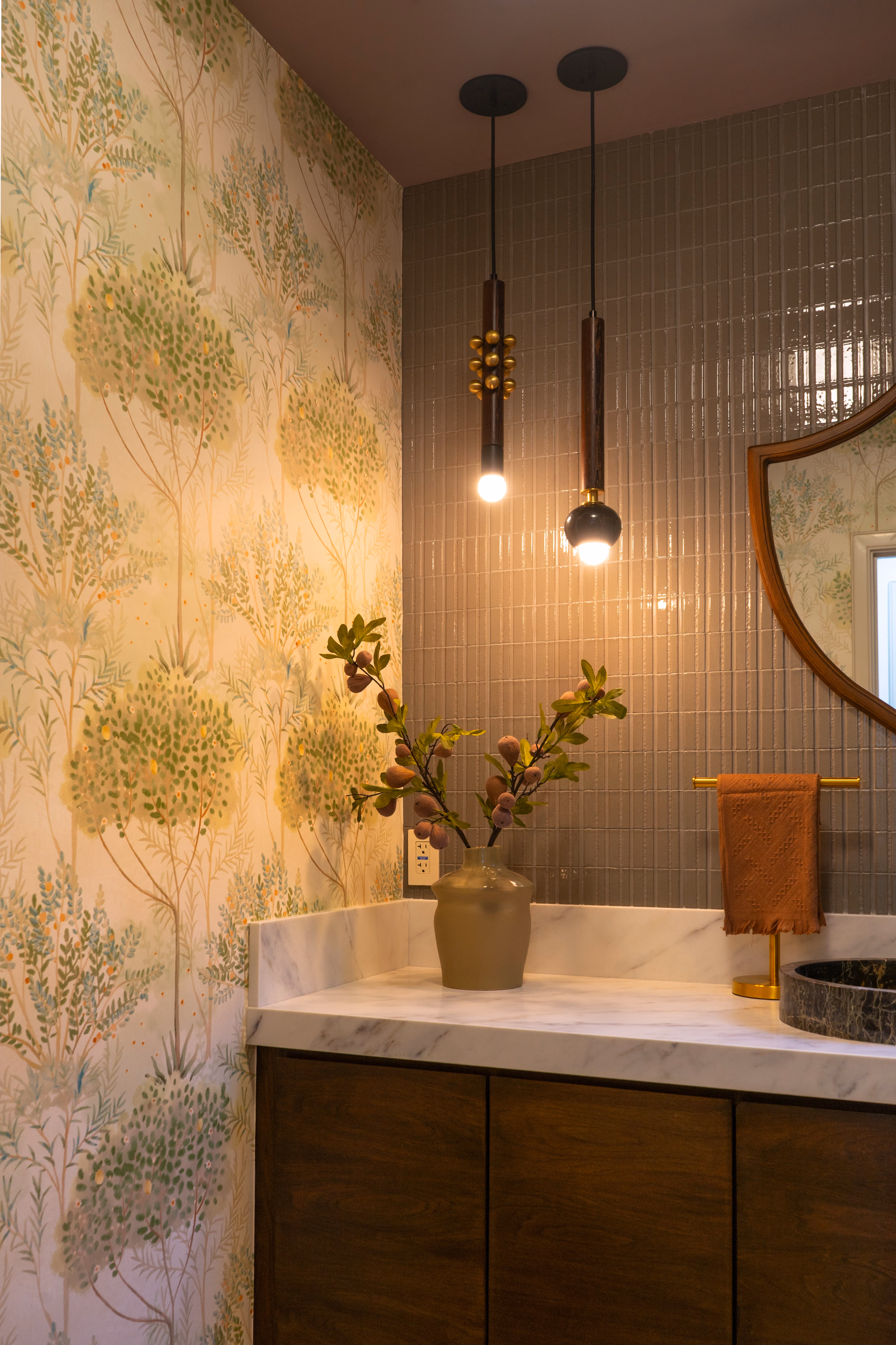 Bathroom corner with floral wallpaper, pendant lights, marble countertop, vase with branches, and towel on gold rack.