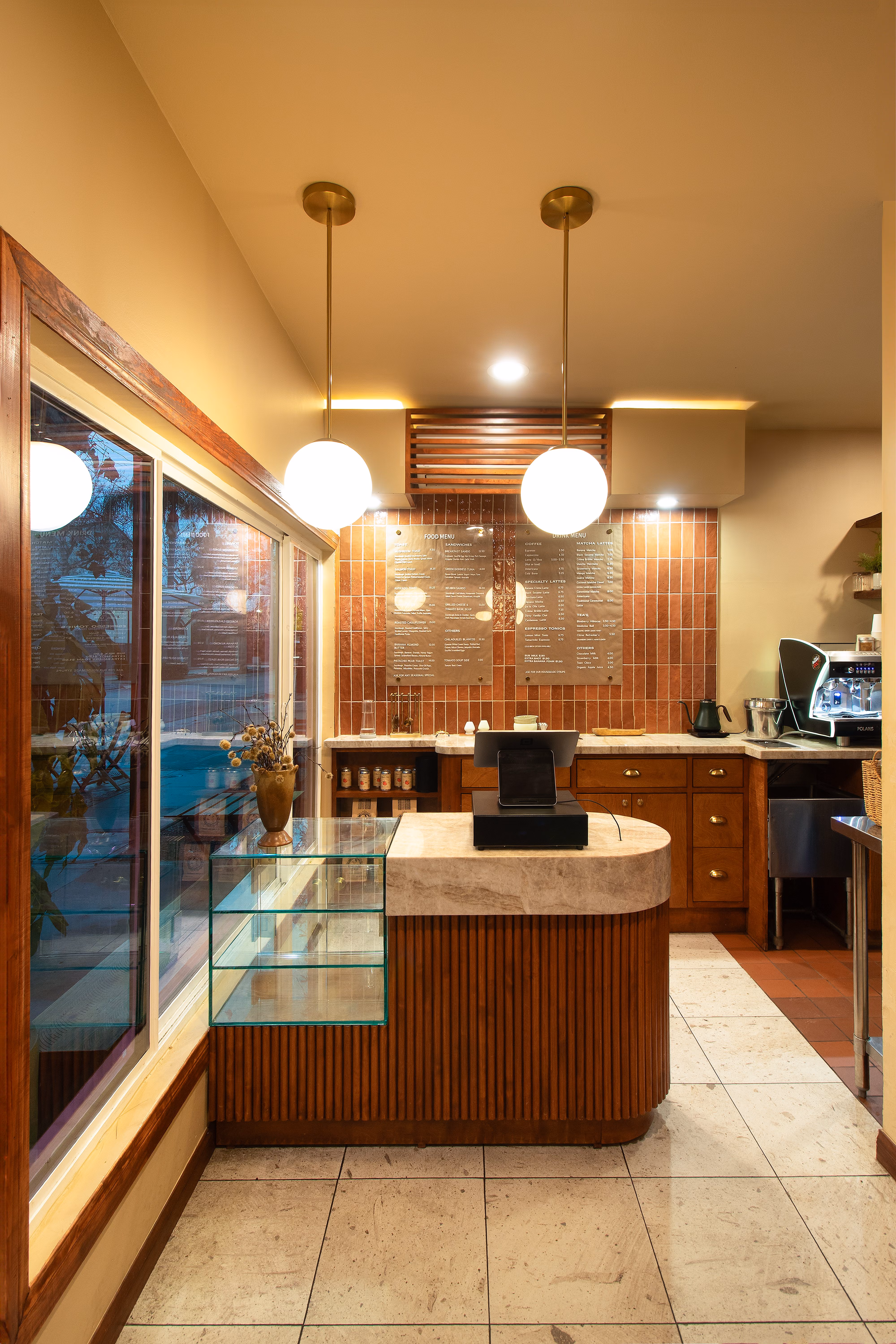 Cozy coffee shop counter with two pendant lights, glass display, cash register, and menu on tiled wall.