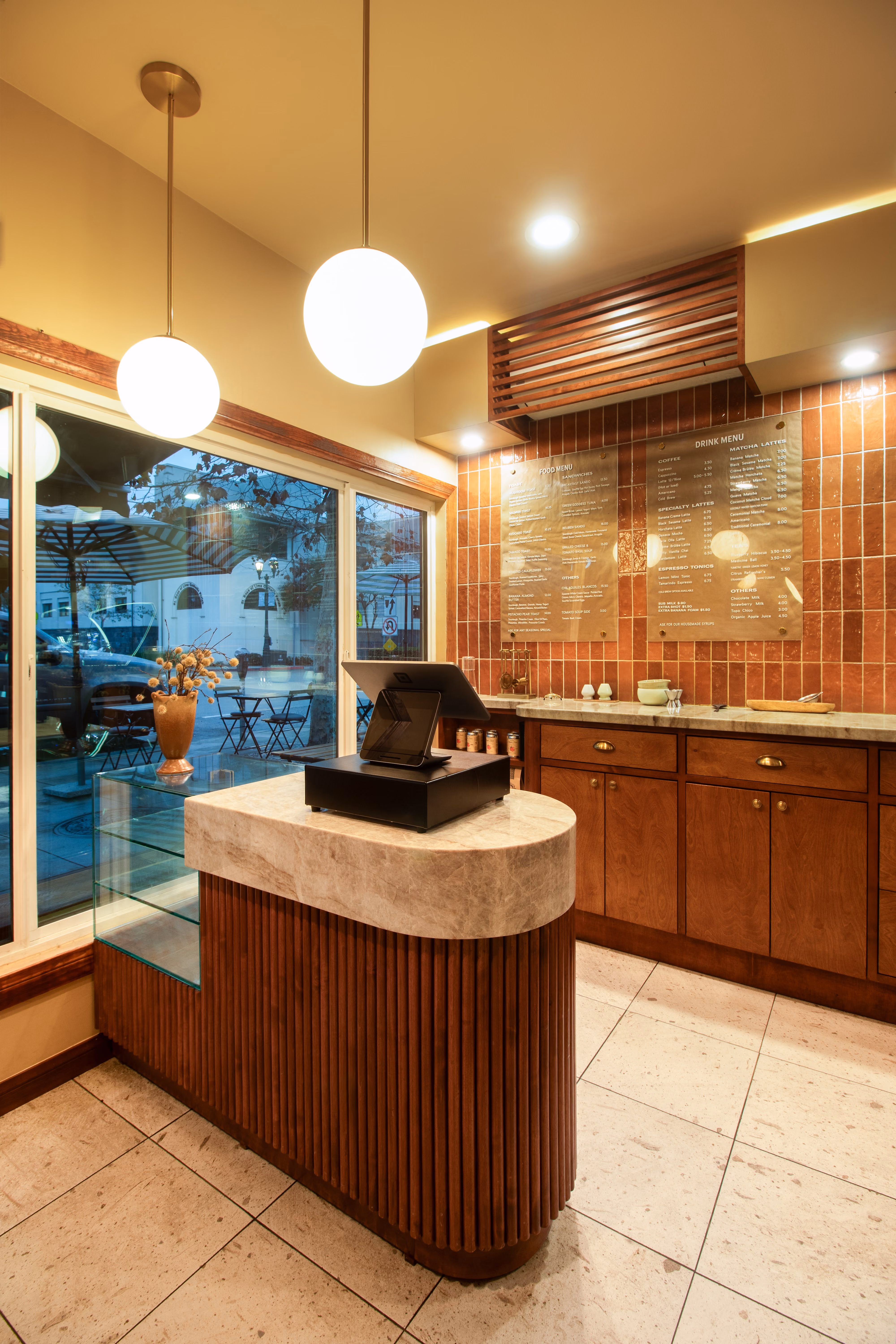 Modern cafe counter with marble top, wooden cabinets, hanging globe lights, and menu boards on tiled wall.